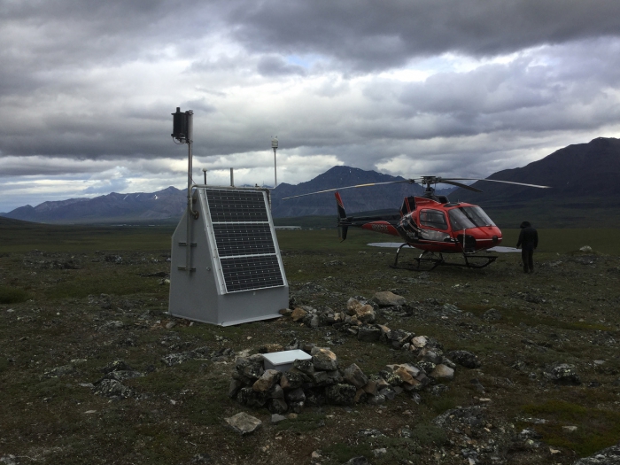 Photo of TA station E22K, located near Anaktuvuk Pass in the Brooks Range, on 18 July 2018. On this service trip, IRIS staff installed two additional lithium batteries and a wind turbine for extra power (the black cylinder on the left). The box in the foreground marks the top of the seismometer borehole. Photo by Max Enders, IRIS